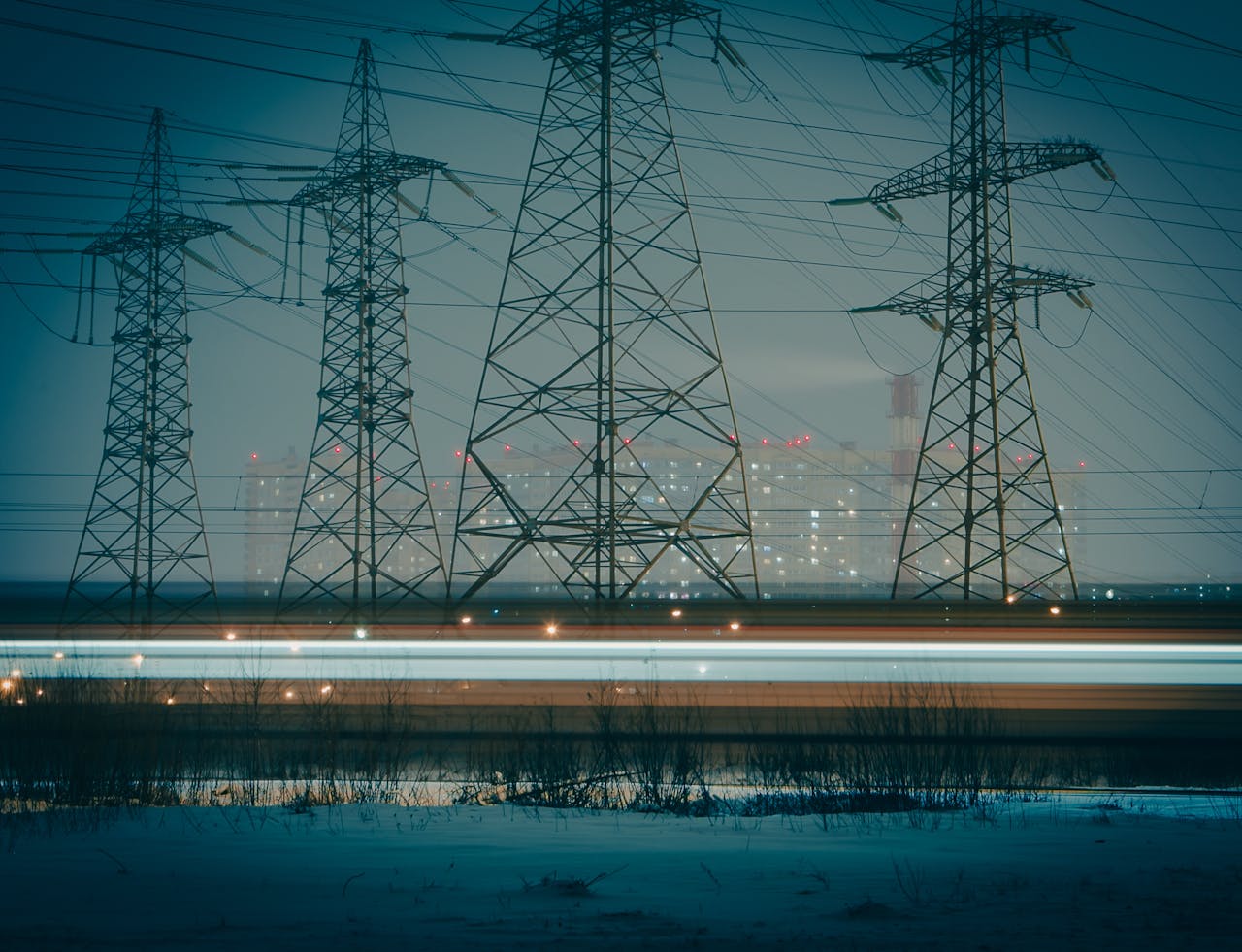 About Moody night photo of electric towers silhouetted against an urban cityscape, highlighting powerful infrastructure.