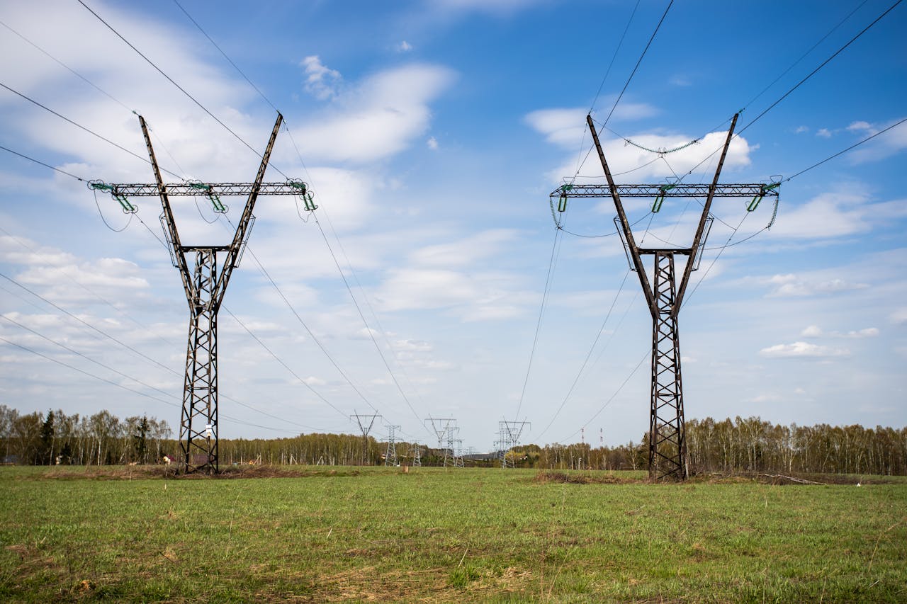 High voltage power lines stretch across a green grass field under a clear blue sky.